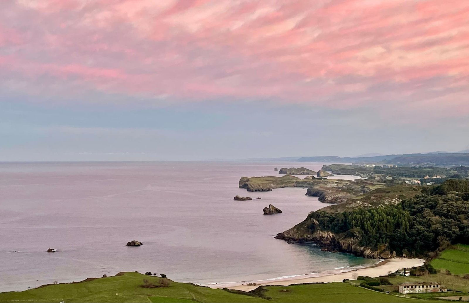 Atardecer en la Playa de Toranda, Niembro (Llanes, Asturias). Panorámica con cielo de tonos rosa, azul y plateado reflejado en el mar en calma, mostrando la ensenada en forma de concha y los acantilados de la costa en el horizonte.