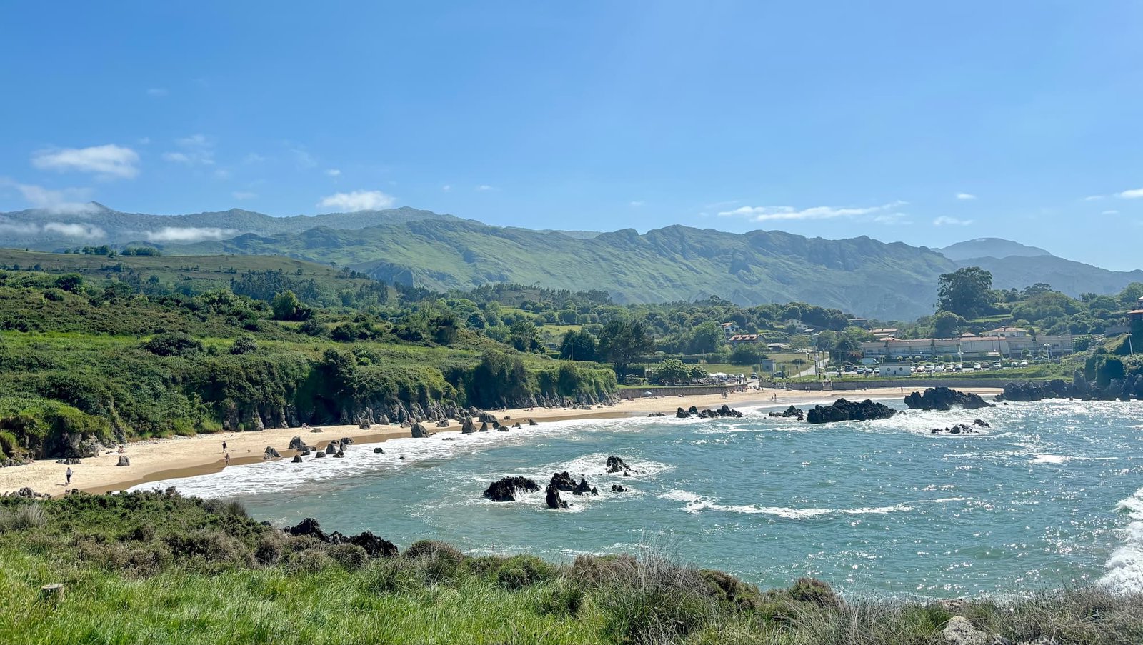 Vista de la playa de Toró en Llanes, Asturias, en un día soleado. Se observa la arena, las rocas, el mar y al fondo las montañas verdes características de la región.