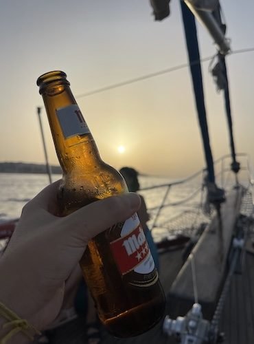 Atardecer en Malta - Mano sosteniendo cerveza fría en la cubierta de un barco, con el mar tranquilo en pleno atardecer