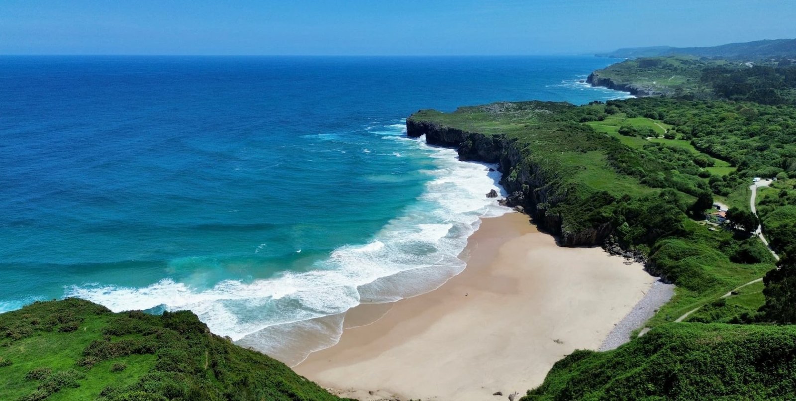 Vista aérea con dron de la playa de Andrín en Llanes, Asturias, mostrando su característica forma de concha perfecta, arena dorada, aguas turquesas y el intenso verde de las praderas que la rodean.
